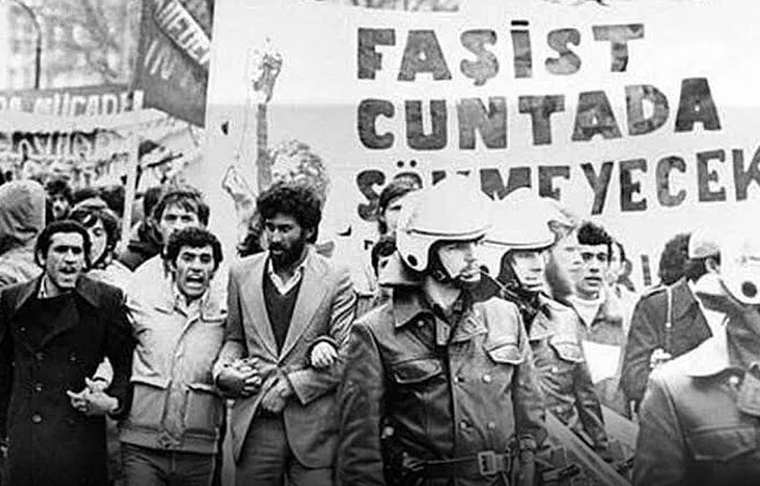 The text on the sign is, "Even the fascist junta will not remove us." This is a protest in Turkiye during the 1980s by socialists. There are military police on the right observing the protest.